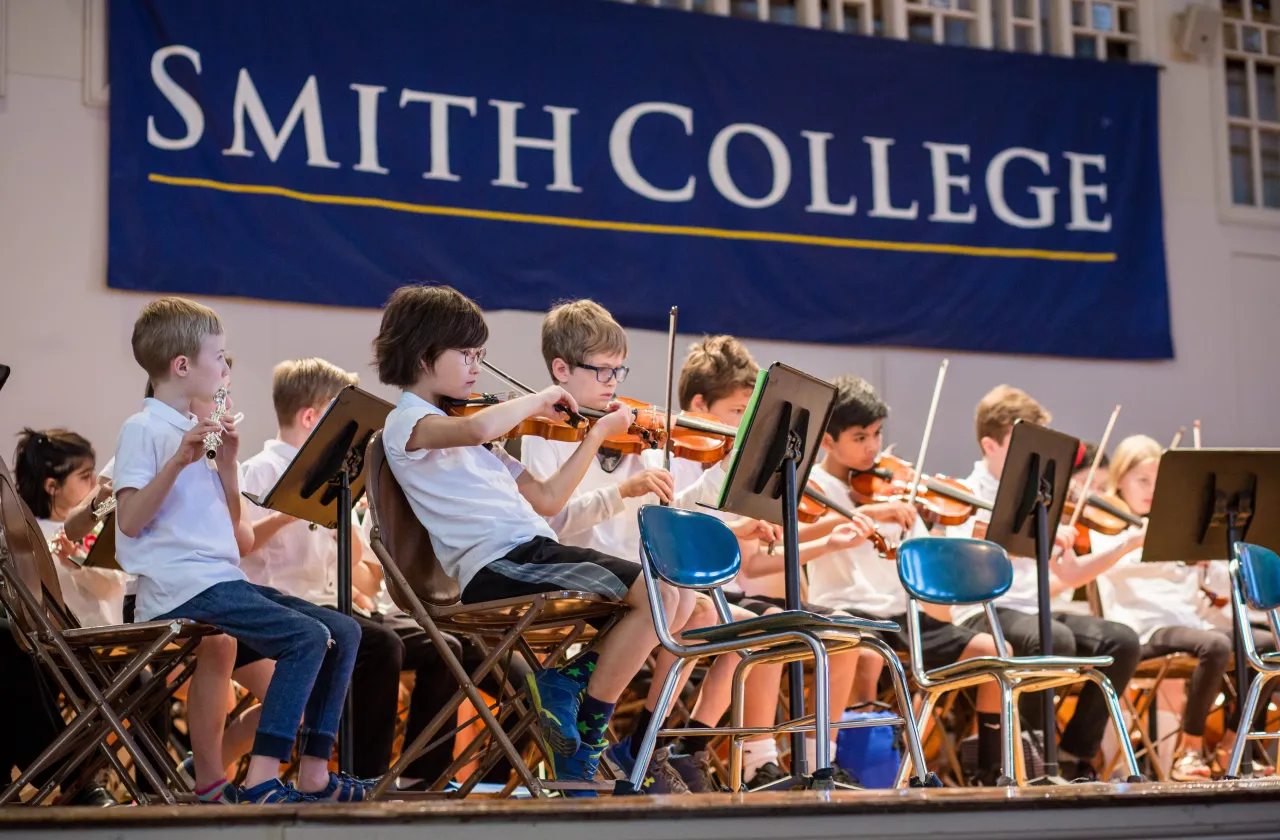 Schoolchildren playing instruments in front of a banner that reads Smith College, in John M. Greene Hall