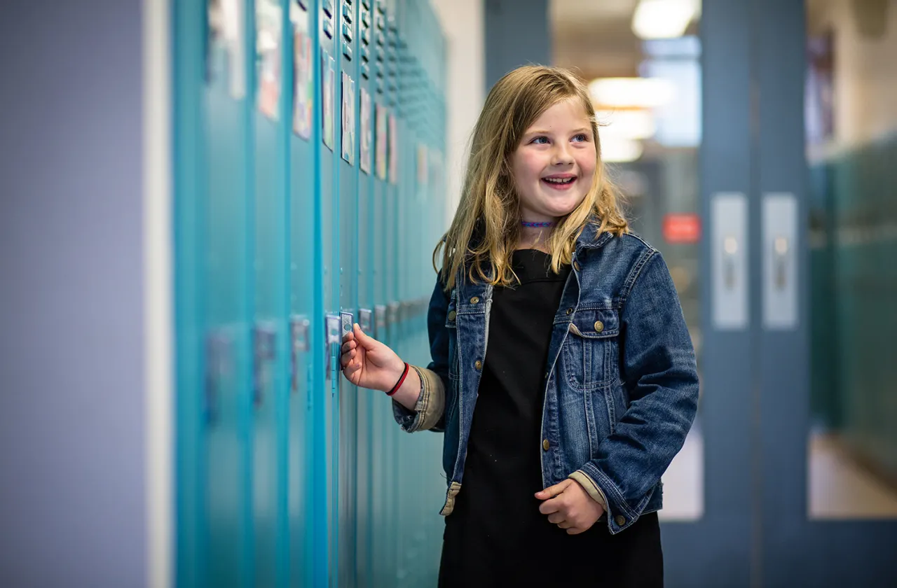 A young girl in a denim jacket standing next to a row of teal lockers, smiling.