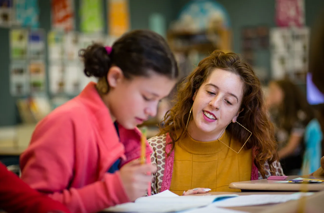 A teacher in a gold shirt talks to a student in a pink jacket.